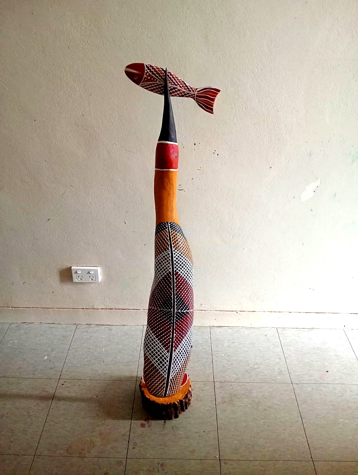 Tall Yolŋu carved bird sculpture balancing a painted fish on its beak